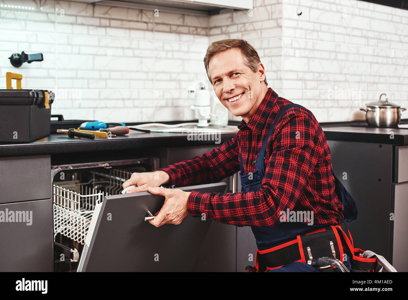 Die professionelle Call. Männliche Techniker in der Nähe von Spülmaschine sitzen mit Schraubendreher in Küche mit Instrumenten und lächelnd Stockfoto