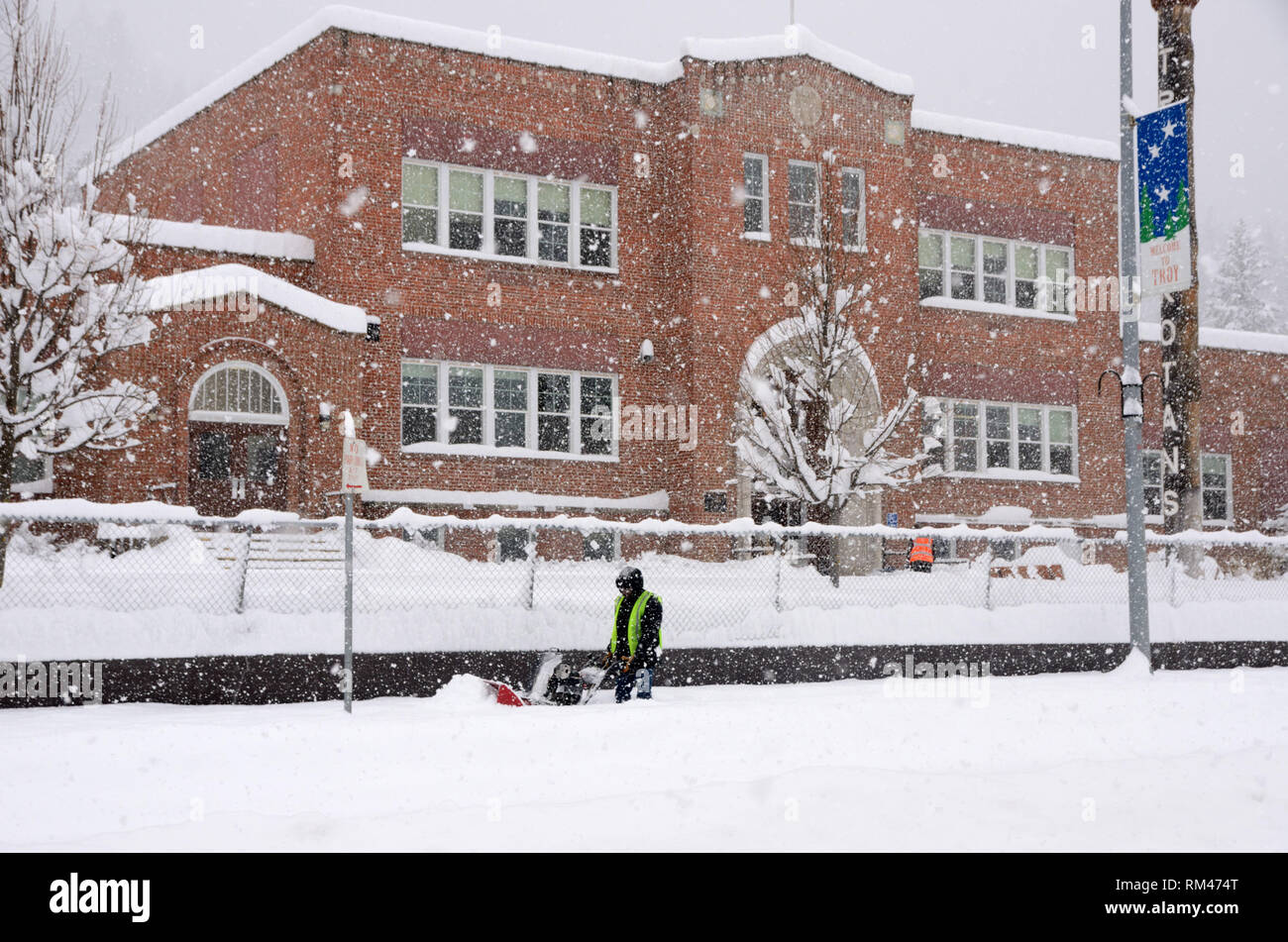 Während eines Schneesturms im Winter arbeitet ein Mitarbeiter vor der öffentlichen Highschool in Troja, Montana, an der Räumung von Bürgersteigen mit einer Schneefräse. Lincoln County, Nordwest-Montana. (Foto: Randy Beacham) Stockfoto