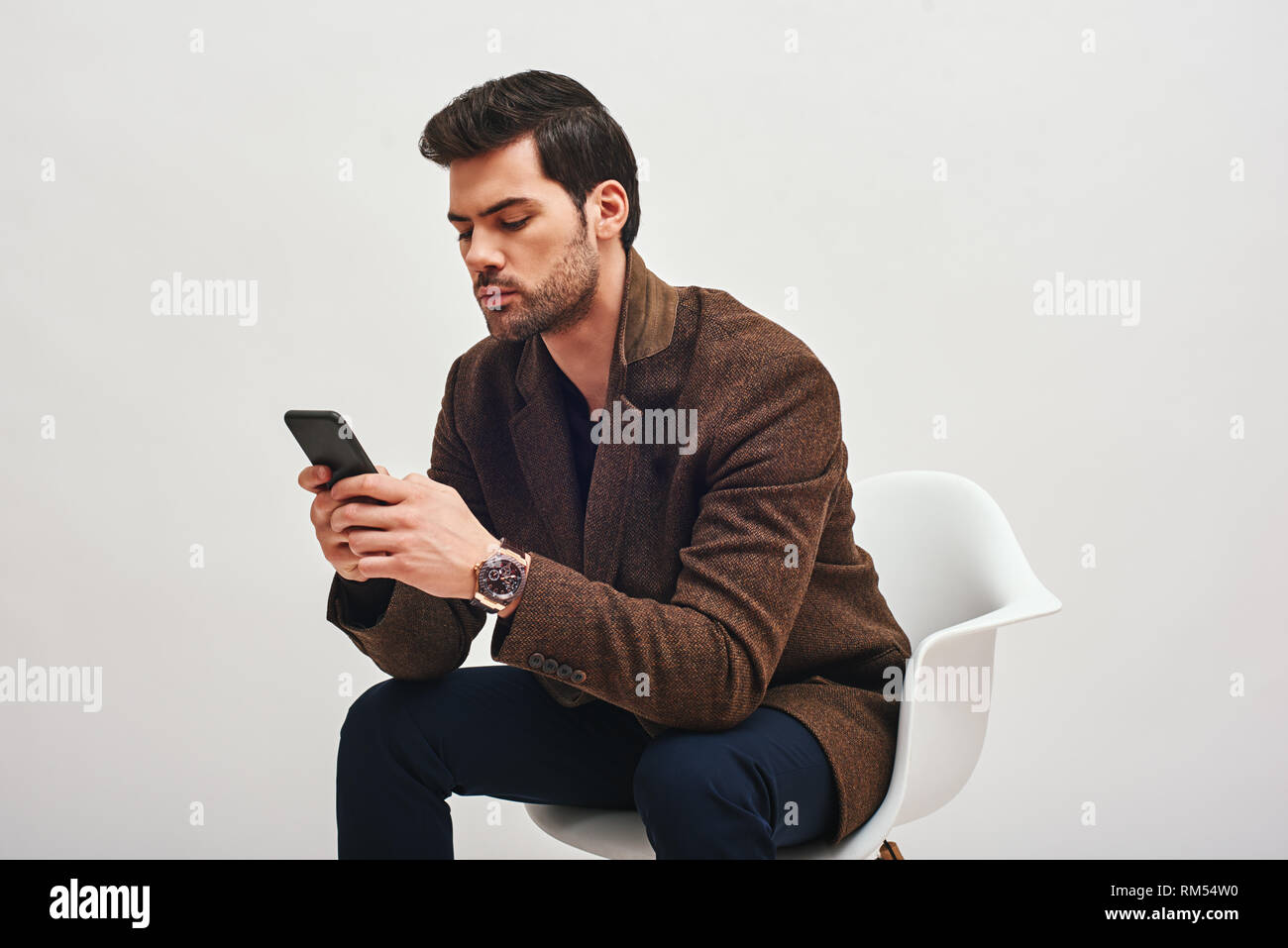 In der Nähe von eleganten dunkelhaarigen Mann trägt blaue Hose und brauner Jacke, mit einer teuren Uhr auf seinem Handgelenk, sitzend auf einem weißen Stuhl und suchen bei seinem Telefon Stockfoto