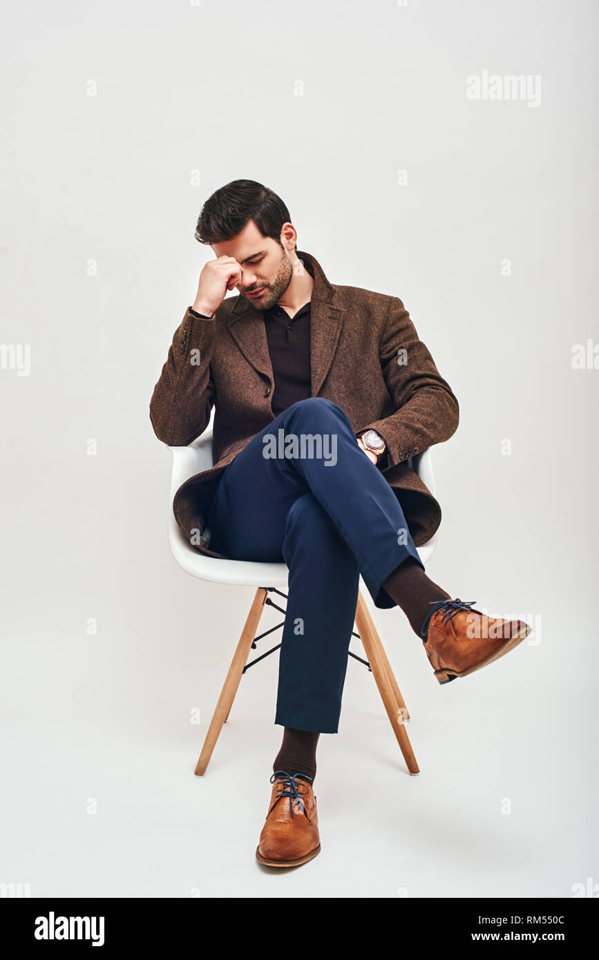 Full-length Portrait von stilvollen dunkelhaarigen Mann trägt blaue Hose und brauner Jacke, sitzen auf einem weißen Stuhl, nach einem harten Tag auf weißem Hintergrund erschöpft Stockfoto