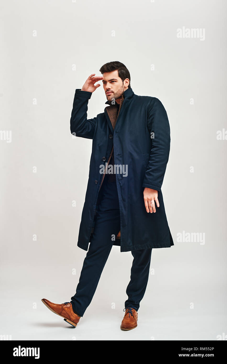 Full-length Portrait von stilvollen dunkelhaarigen Mann trägt blaue Hose, braun jacke und Blue Coat stehen und auf der Suche nach Etwas auf weißem Hintergrund Stockfoto