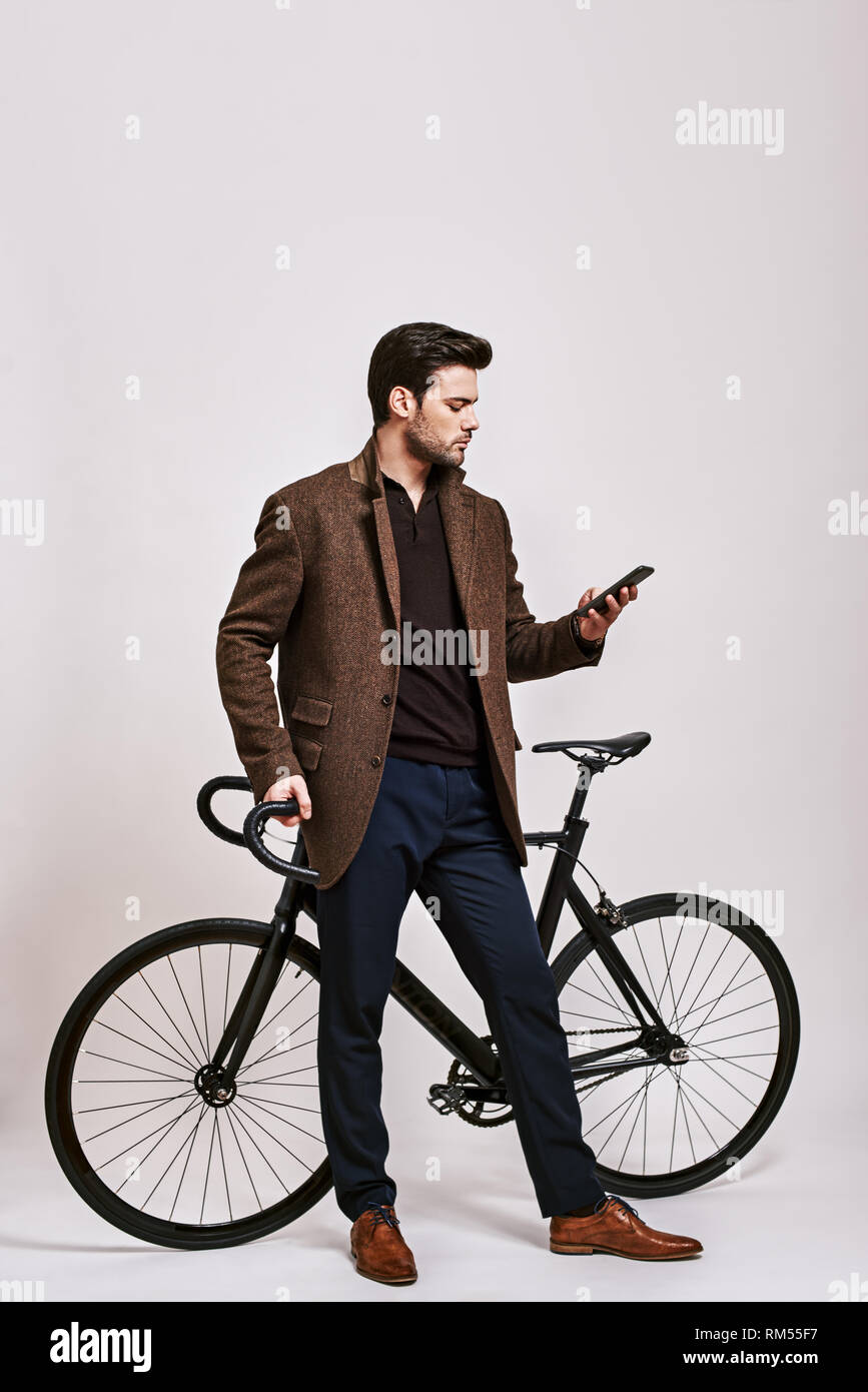 Full-length Portrait von stilvollen dunkelhaarigen Mann trägt blaue Hose, braun jacke mit schwarzen Fahrrad und suchen bei seinem Telefon isoliert über grauer Hintergrund Stockfoto
