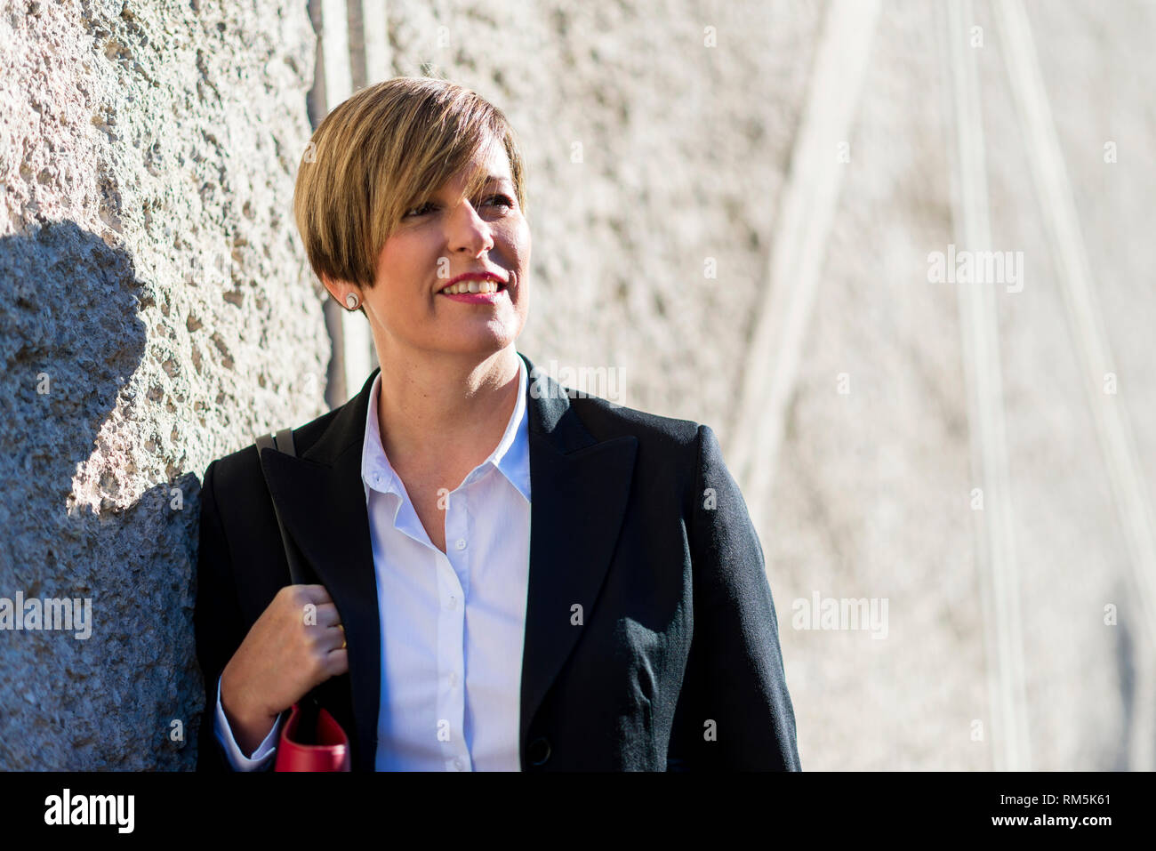 Vorderansicht des eleganten lächelnd business Frau lehnte sich auf einem Gebäude, Wand, während das Wegsehen Tagträumen im Freien an einem sonnigen Tag Stockfoto