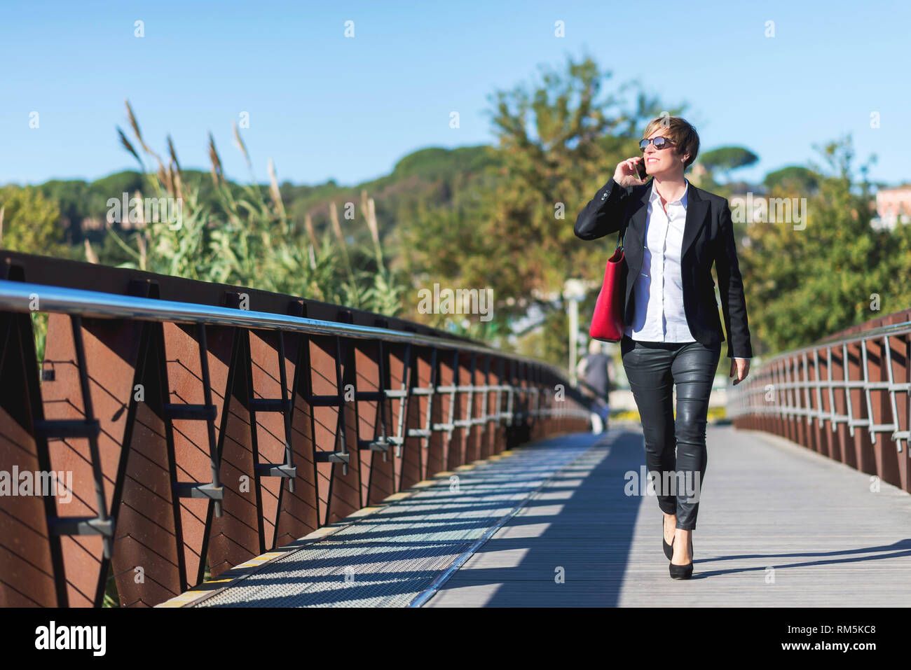 Vorderansicht des eleganten Business Frau mit einem roten Tasche zu Fuß auf den Weg, während Sie ein Mobiltelefon und Wegsehen im Freien Stockfoto