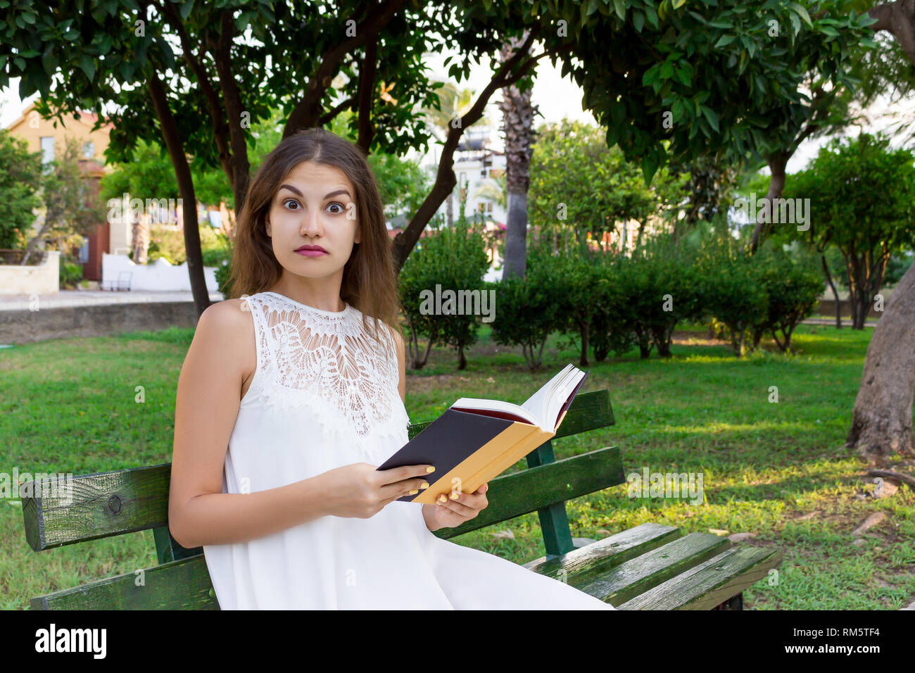 Closeup Portrait von einem schönen Mädchen überrascht mit weit geöffneten Augen, die beim Lesen eines Buches auf der Bank im Park. Menschliche emotion Gesichtsausdruck f Stockfoto