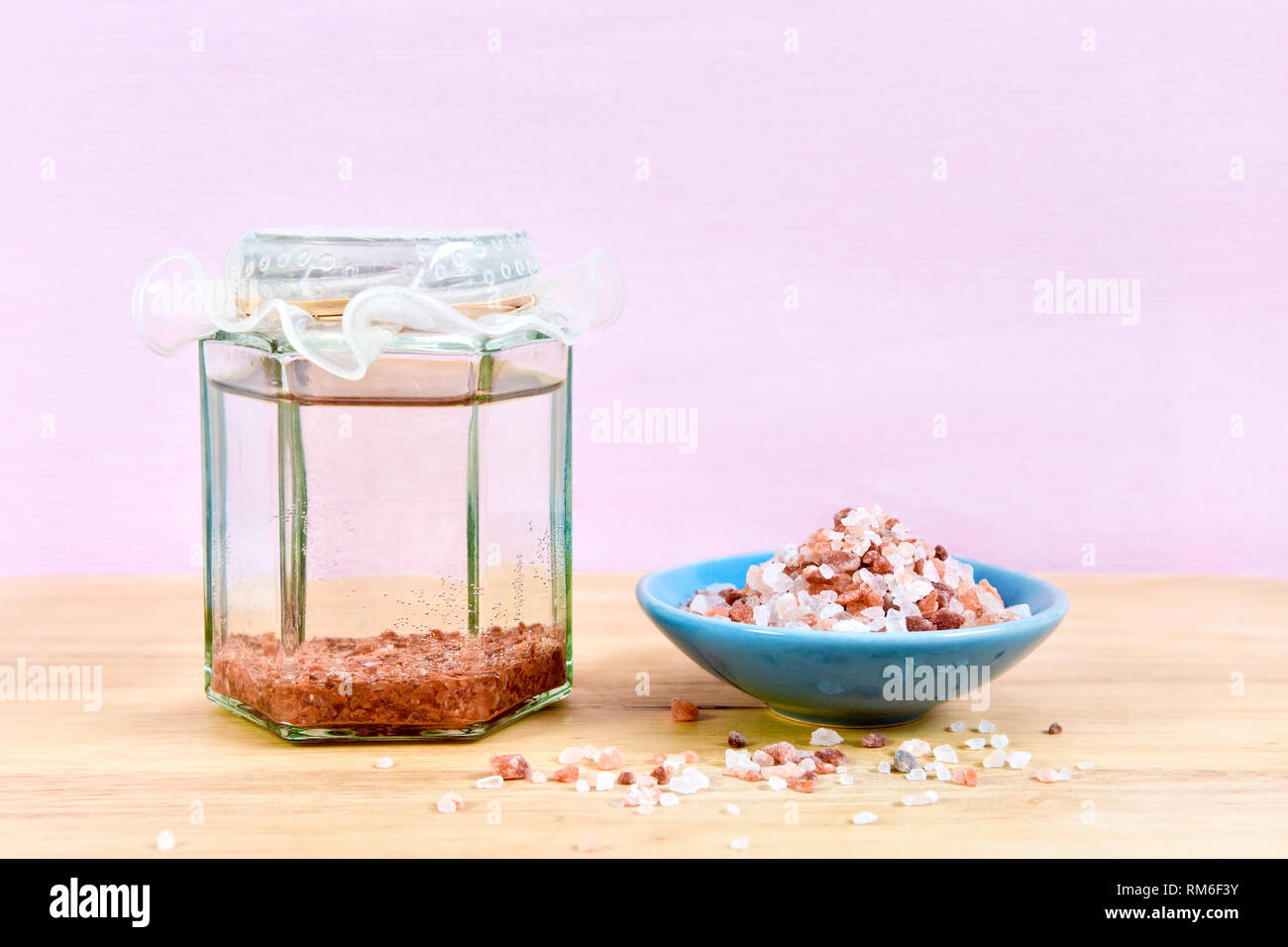Himalaya Salz Sole, Sole oder Wasser, in Glas Glas mit einer Schüssel mit Himalaya Salz, auf einer hölzernen Schneidebrett, gegen einen rosa Hintergrund Stockfoto