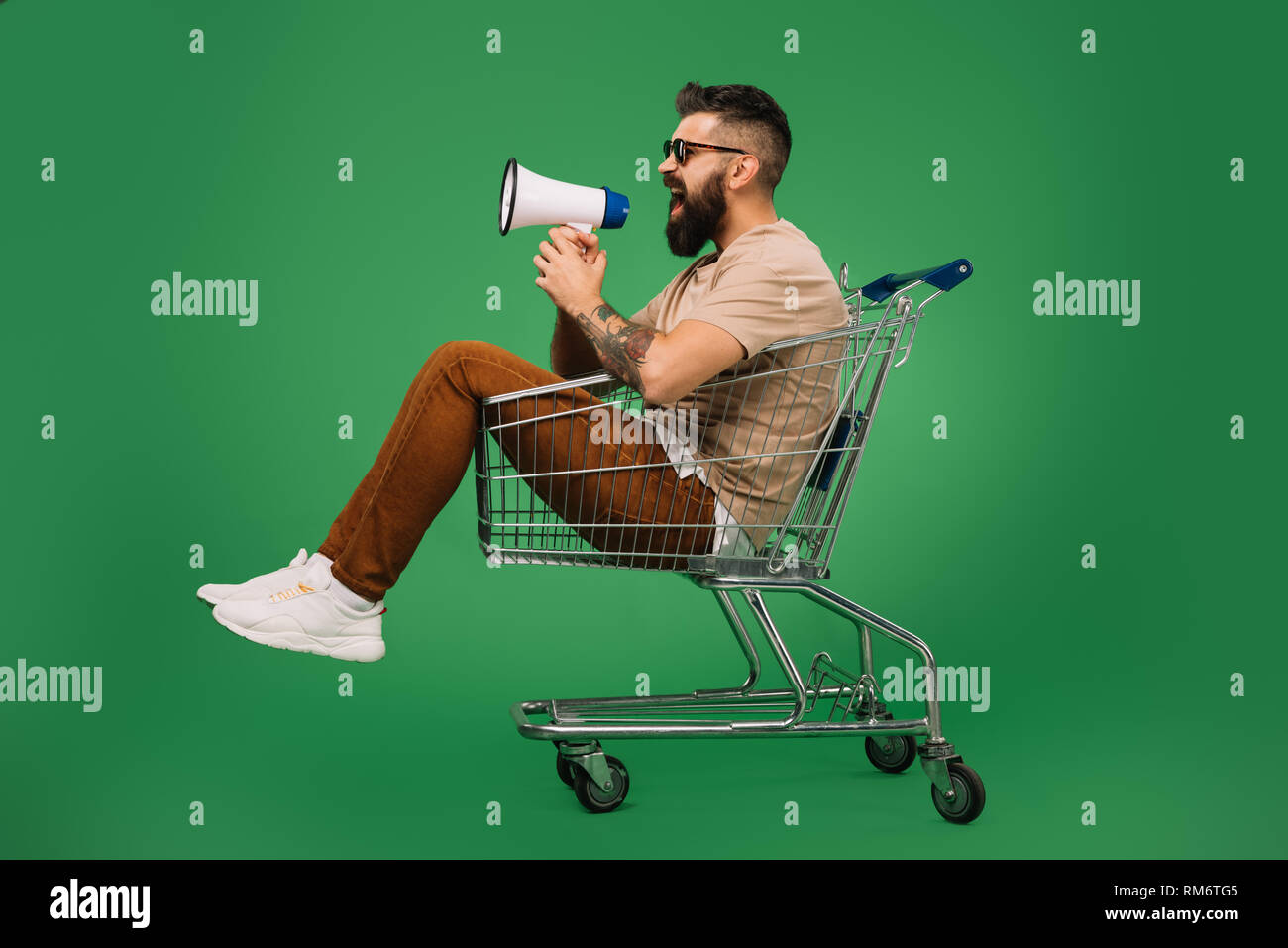 Bärtiger Mann schreit in Megaphon während in Warenkorb sitzt isoliert auf Grün Stockfoto