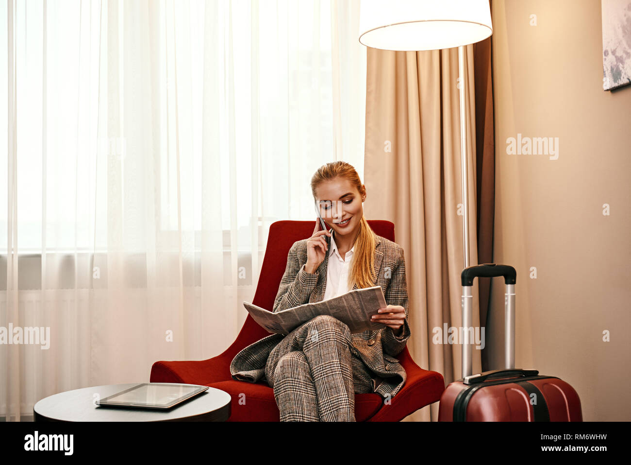 Business Call im Hotel Zimmer in der Geschäftsreise. Junge und stilvolle Geschäftsfrau mit Koffer und Smartphone sitzt auf einem Sofa im Hotel Zimmer. Digitale tablet am Tisch Stockfoto