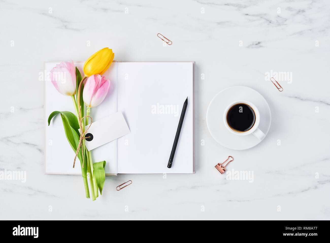Schreibtisch mit Kaffeetasse, Büroklammern, Bleistift, rosa und gelbe Tulpen mit Tag für Name oder Nachricht auf offenen Tagebuch über weißem Marmor Hintergrund. Flach Stockfoto