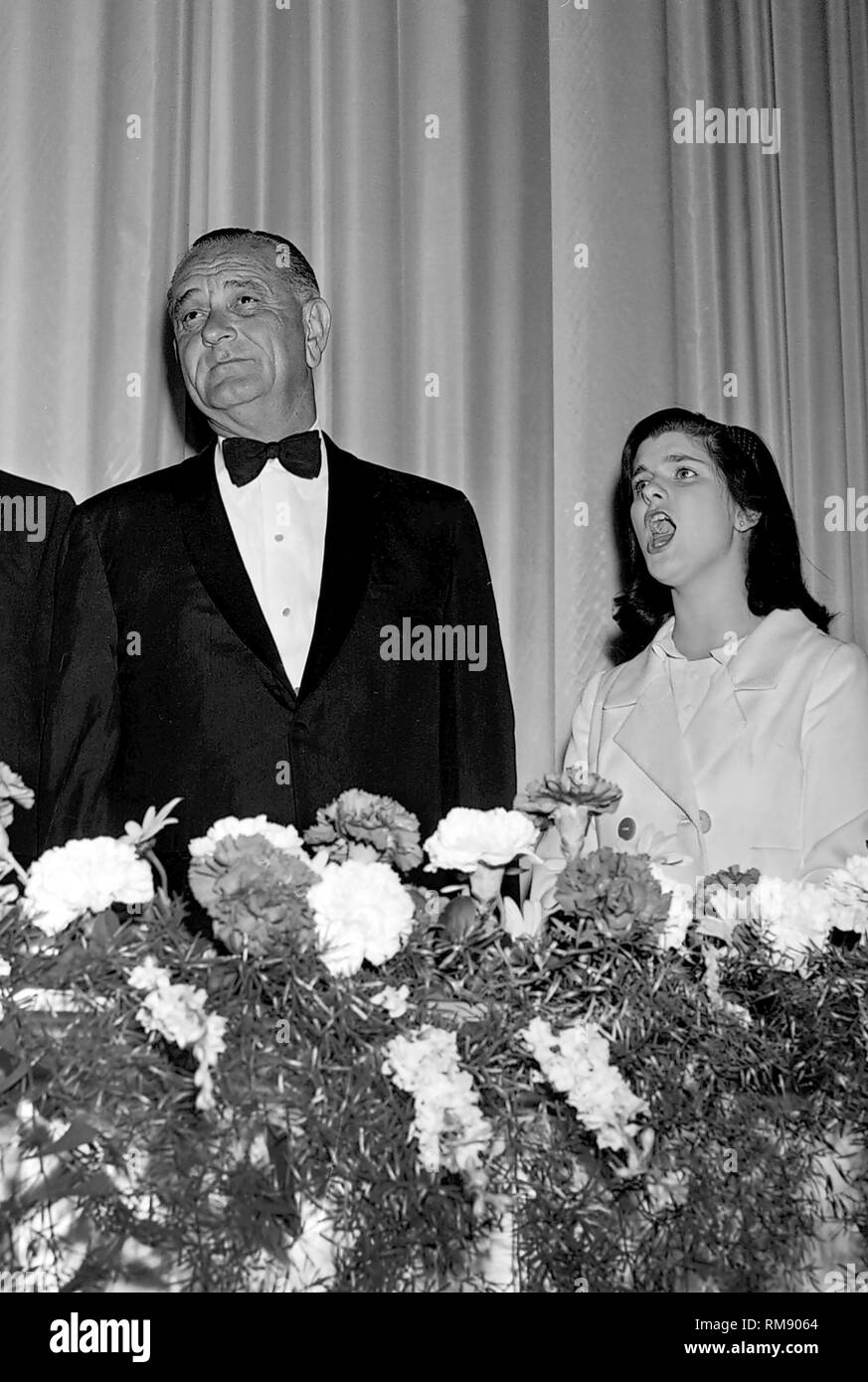 Präsident Lyndon B. Johnson steht aufmerksam, während eine junge Frau die Nationalhymne neben ihm bei einem Abendessen in das Conrad Hilton Hotel in Chicago singt, Ca.1964. Stockfoto