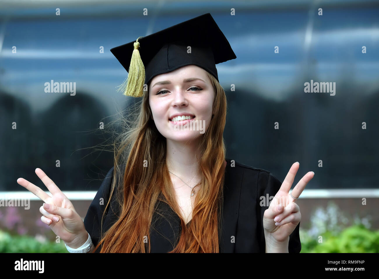 Gerne Absolvent junges Mädchen mit roten Haaren in schwarzem Mantel und Square akademischen Gap im Campus der Universität bekleidet zeigt Sieg Zeichen Stockfoto