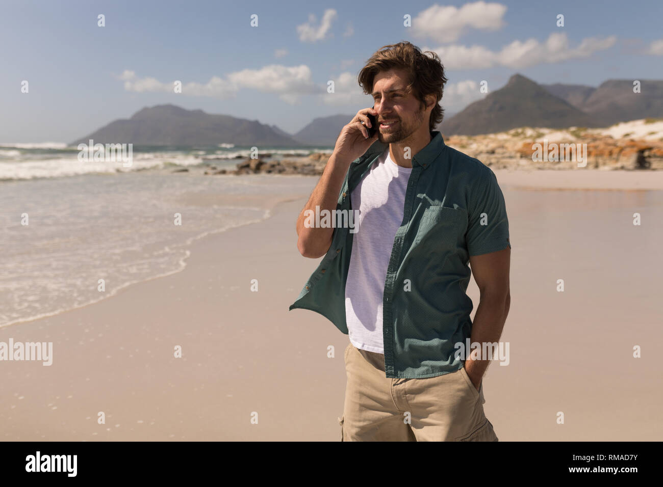 Junger Mann mit der Hand in der Tasche Gespräch am Handy am Strand Stockfoto