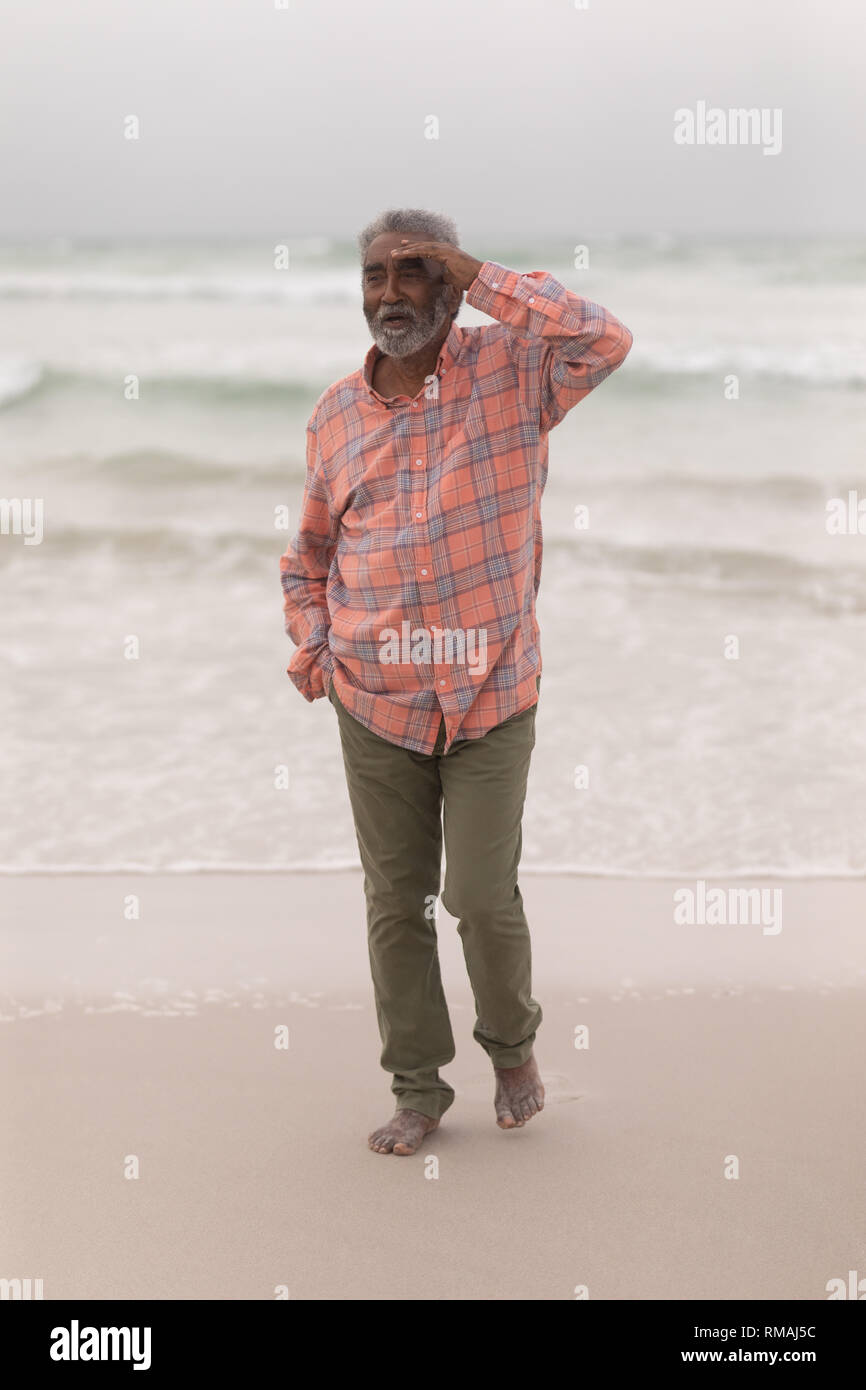 Älterer Mann mit Abschirmung Augen und Hand in der Tasche zu Fuß am Strand Stockfoto