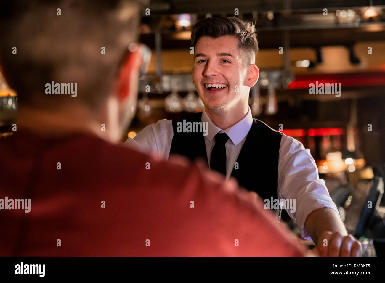 Blick über die Schulter eines jungen männlichen Barkeeper, Lachen ist und im Gespräch mit einem Kunden an der Bar. Stockfoto