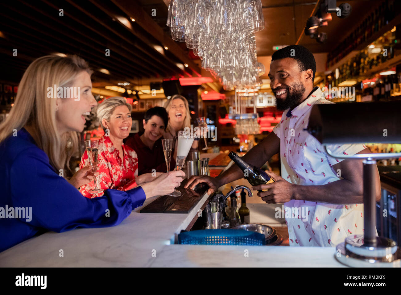 Kleine Gruppe von reifen Frauen genießen eine Nacht, sitzen an der Bar. Sie werden lachen mit dem Barkeeper, der hält eine Flasche Prosecco. Stockfoto