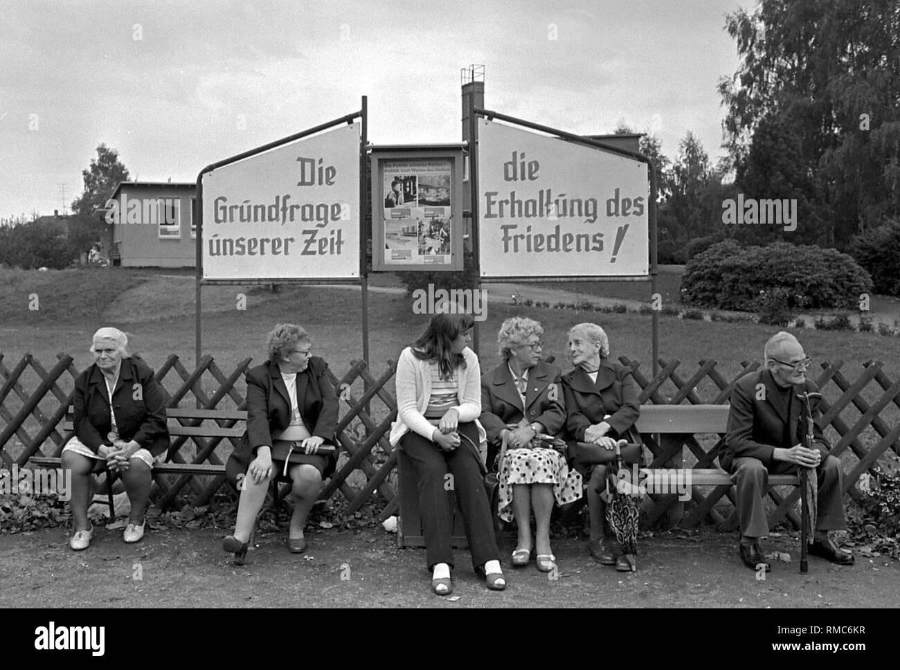 Frauen und ein Mann sitzen und auf zwei Bänke in einer Stadt in der DDR sprechen, hinter Ihnen sind zwei sozialistische Propaganda Plakate mit der Aufschrift "Die grundlegende Frage unserer Zeit - die Erhaltung des Friedens". Stockfoto