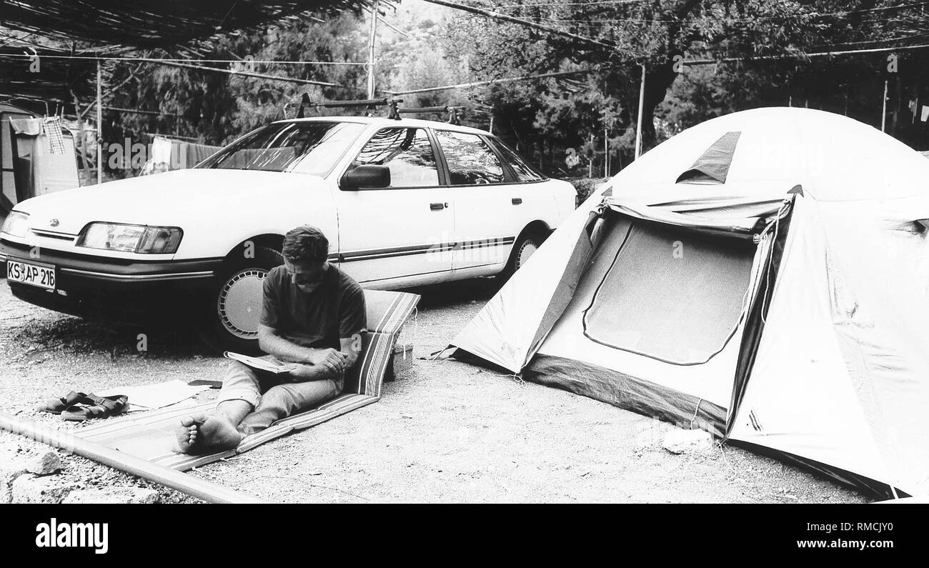 Ein Mann sitzt auf einer Luftmatratze zwischen sein Auto und seine Hütte und liest eine Zeitschrift auf einem Campingplatz an der Lefka Beach (in der Nähe von nauplia). Stockfoto