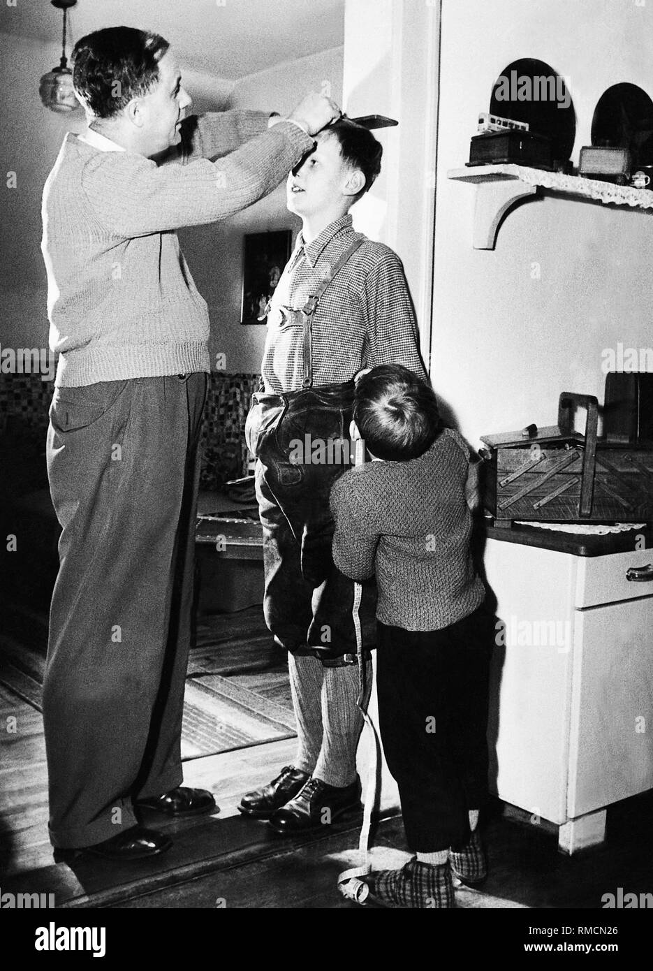 Ein Vater misst die Höhe seines älteren Sohnes. Er markiert die Höhe auf dem Türrahmen mit einem Bleistift, der jüngere Sohn das Maßband hält. Stockfoto