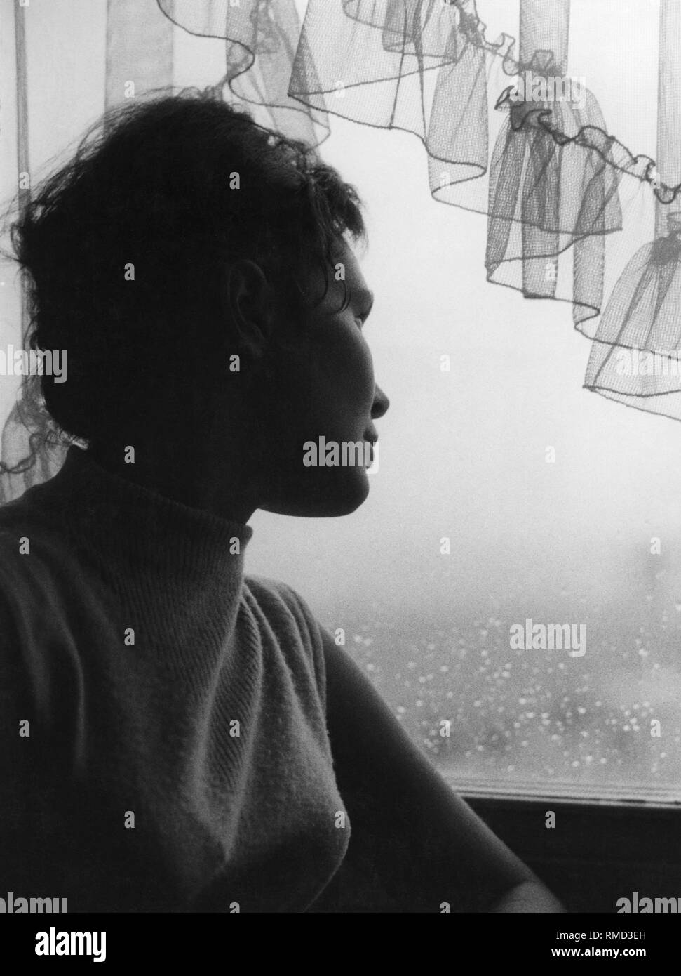 Junge Frau schaut verträumt durch einen regnerischen Fenster, 50 s Stockfoto