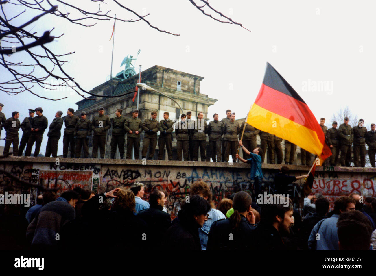 Grenztruppen stand in einer Reihe auf der Berliner Mauer vor dem Brandenburger Tor. Sie wachen über eine Menschenmenge vor der Wand. Ein Mann weht die deutsche Fahne. Stockfoto
