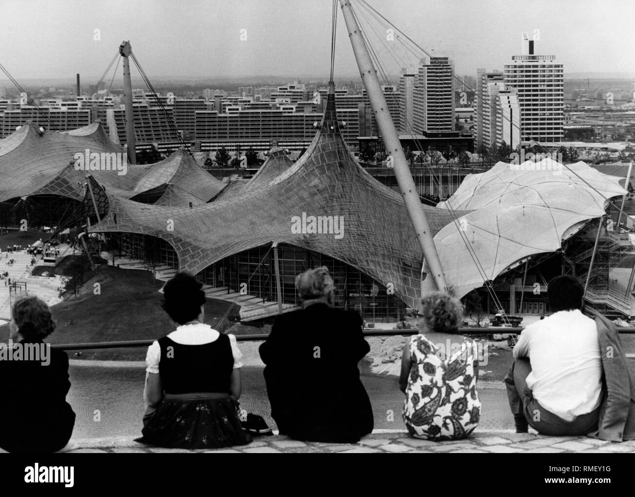 Vier Touristen aus den Münchner Olympiaberg auf dem Olympiagelände in München, wo die olympischen Sommerspiele 1972 statt. Unter ihnen der Olympiasee See, links das Theatron, geradeaus die Olympia-Schwimmhalle. Im Hintergrund das Olympische Dorf. Stockfoto