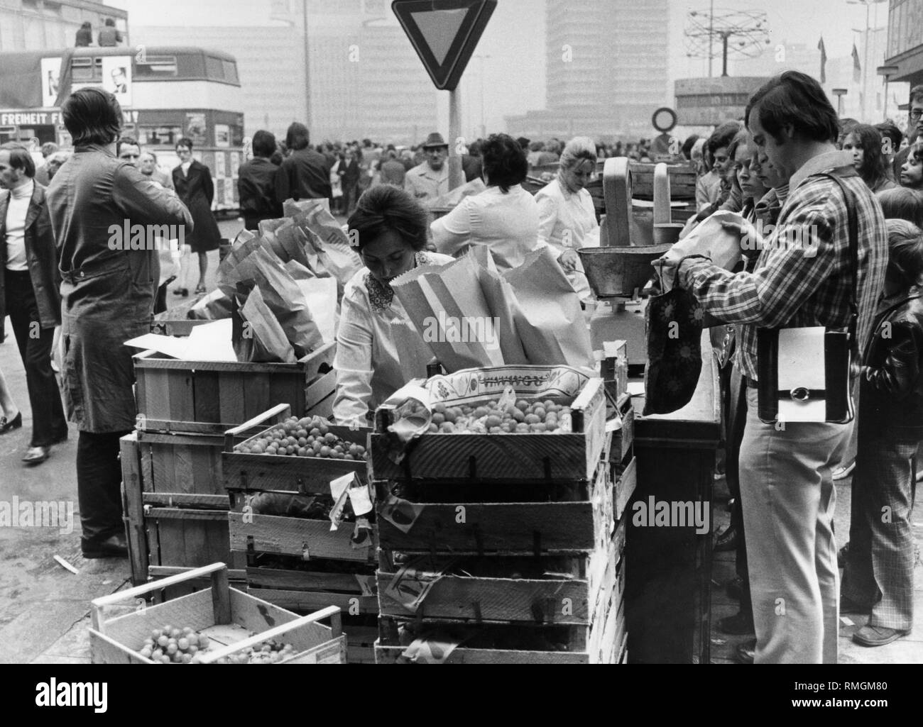 Eine Verkäuferin setzt Trauben in eine Tasche an einem Obststand auf dem Alexanderplatz in Ost-Berlin für den Kunden. Rechts im Hintergrund die Urania Weltzeituhr. Undatiertes Foto. Stockfoto