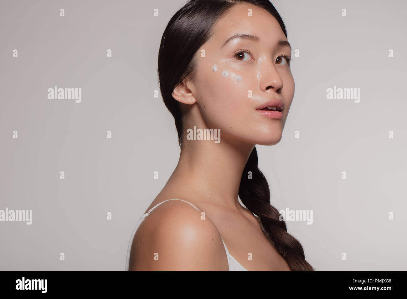Asiatische Frau Modell mit Feuchtigkeitscreme Creme im Gesicht über beigen Hintergrund. Junge Frau mit kosmetischen Creme auf die eine Wange an Kamera schaut. Stockfoto