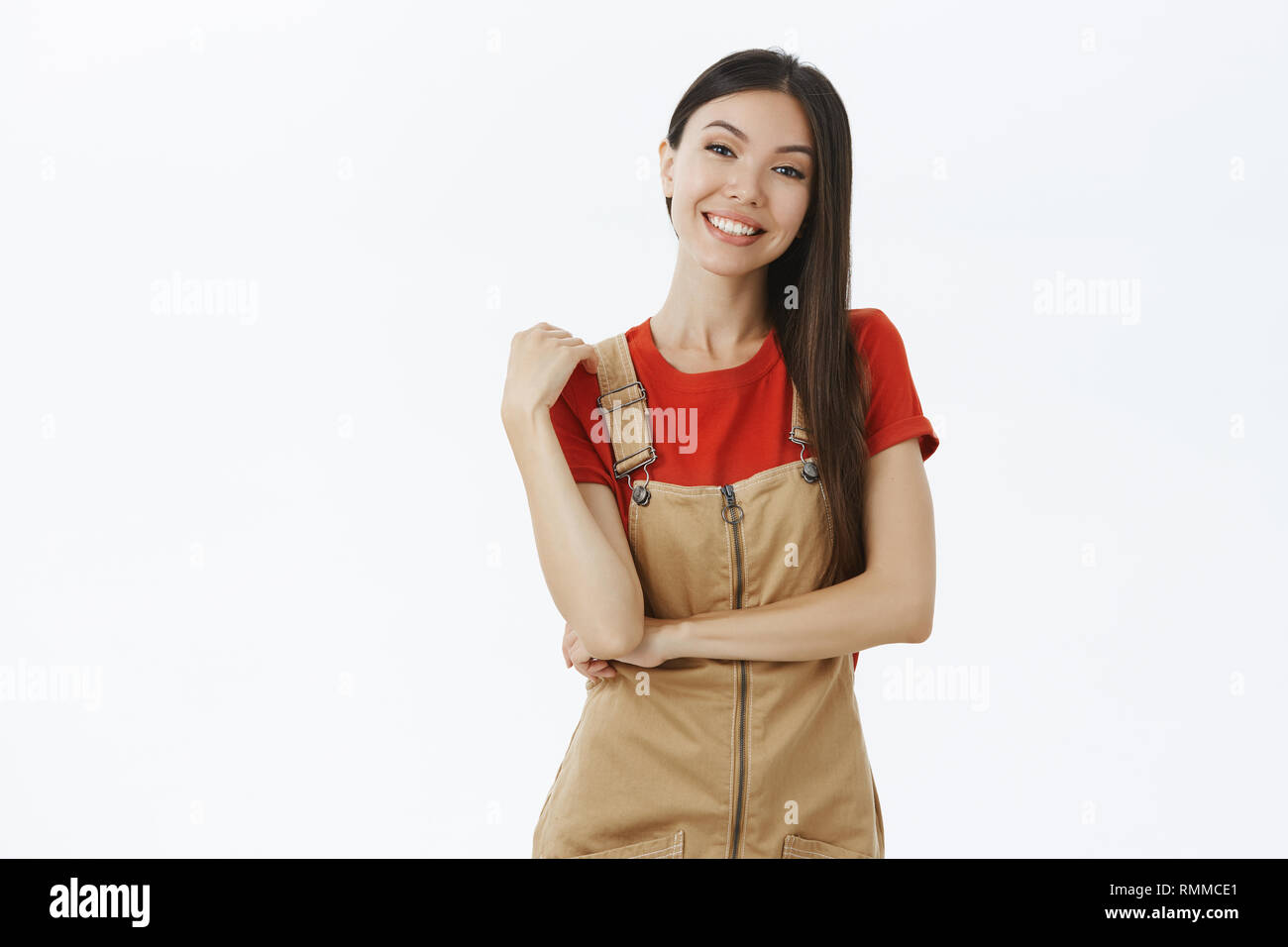 Portrait von erfreut, charismatischen und Ausgehende asiatische Frau mit dunklen Haaren in süßen braunen Overalls berühren Schulter, während Lächeln hören von Interesse Stockfoto