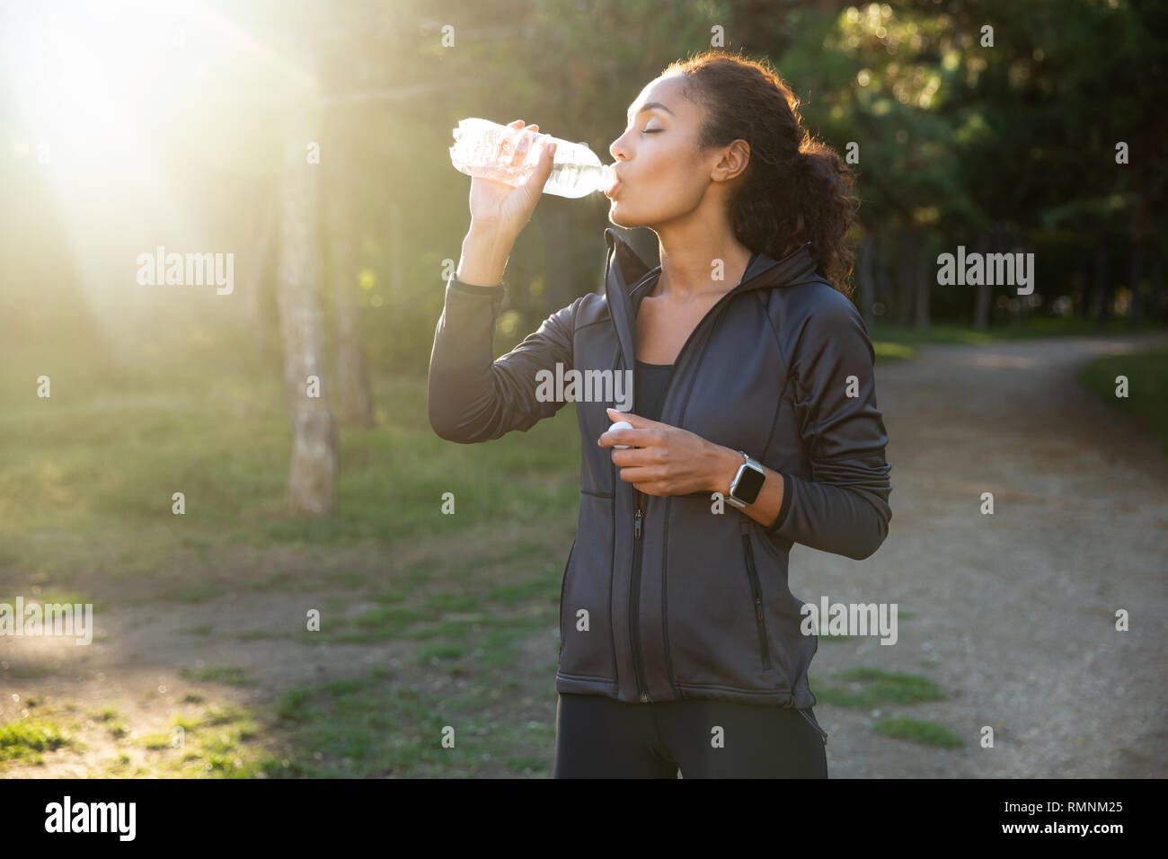 Bild der athletische Frau 20s mit schwarzen Trainingsanzug Trinkwasser bei einem Spaziergang durch den Green Park Stockfoto