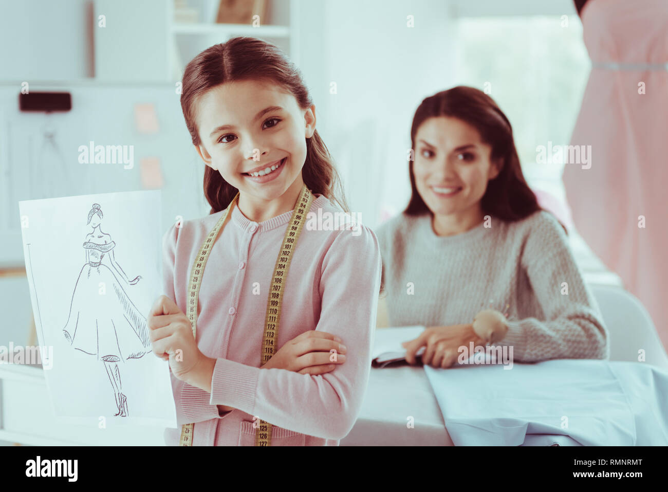 Brillante Arbeit. Portrait von kleinen Mädchen, dass eine Modellskizze und halten die Hände gekreuzt Position, während ihre Mutter besetzt mit Stoff Stockfoto