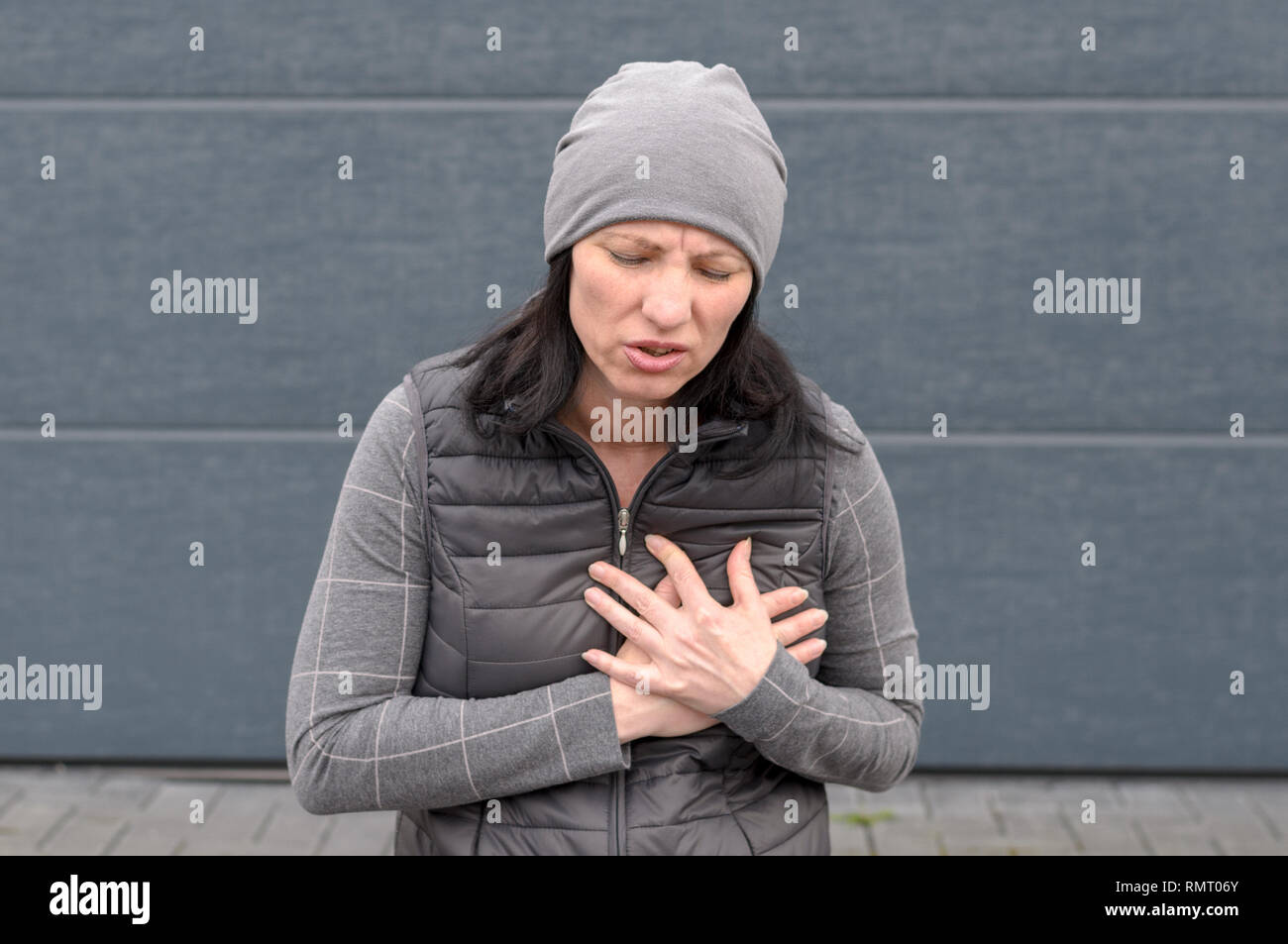 Frau, die draußen in einem grauen Winter Outfit umklammerte ihre Brust mit beiden Händen und Grimassen in den Schmerz in ein Konzept von einem Herzinfarkt oder Herzstillstand pr Stockfoto