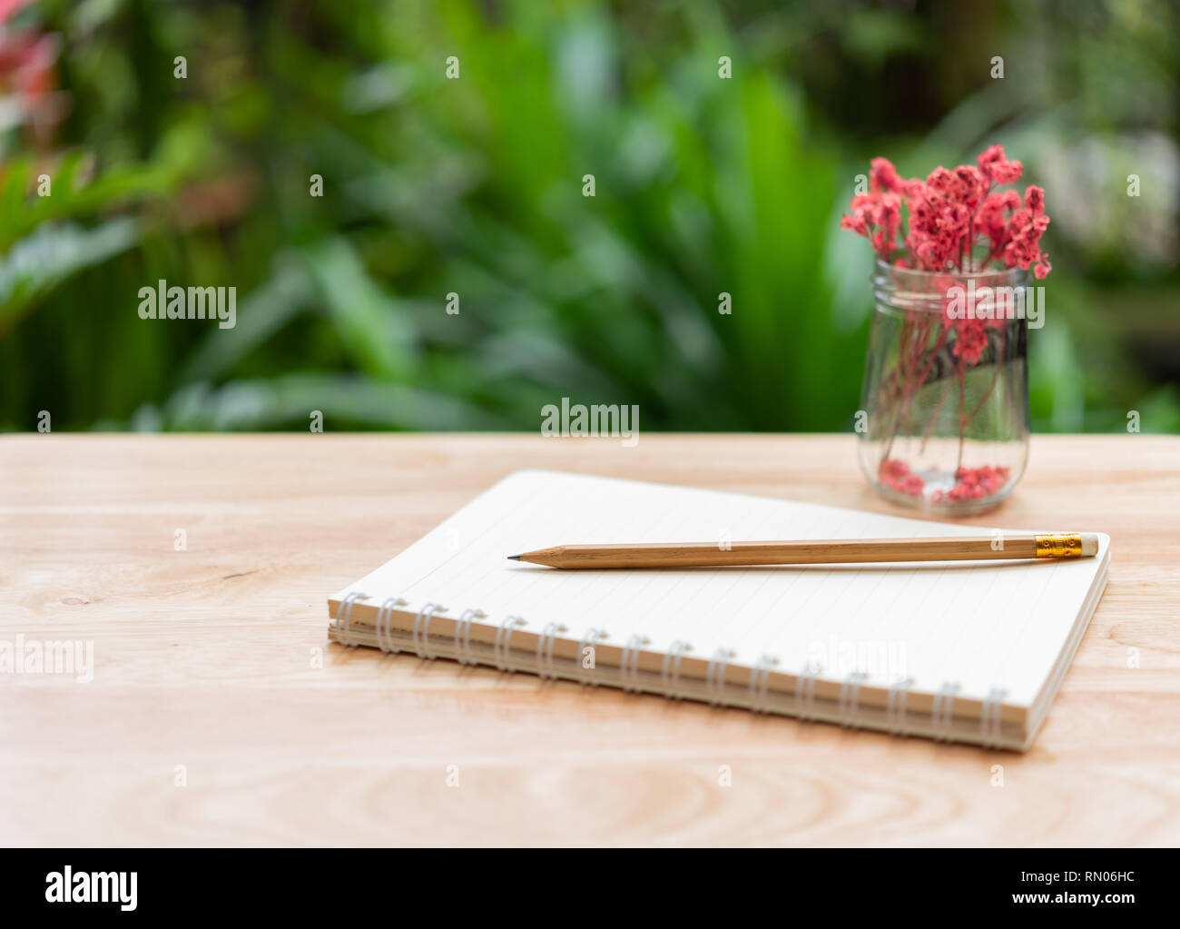 Notebook, braun Bleistift und schöne rote getrocknete Blumen im Glas Vase auf Holz Tisch mit frischen grünen Garten Hintergrund, outdoor Arbeitsbereich Stockfoto