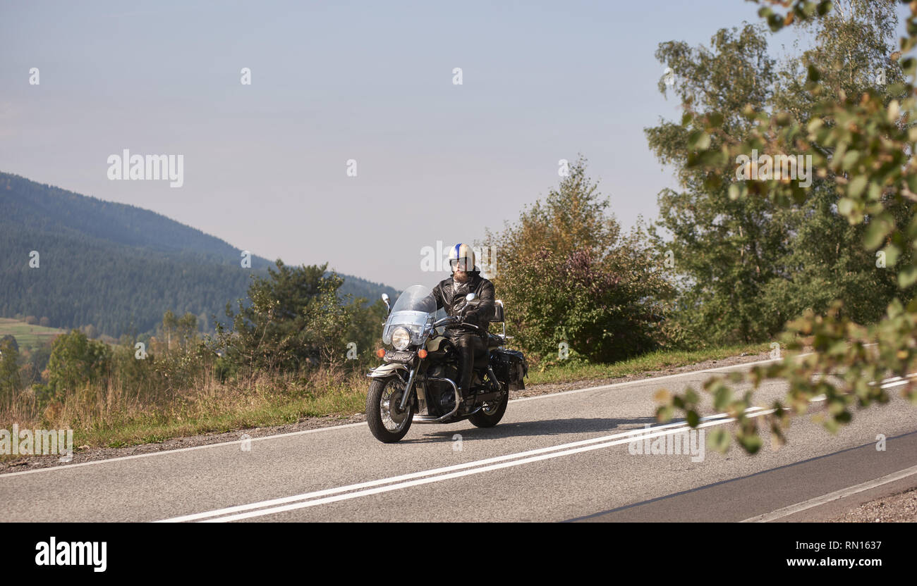 Bärtige Biker in Helm mit Sonnenbrille und schwarzem Leder kleidung reiten Cruiser leistungsstarke Motorrad, sonnigen Asphaltstraße auf hellen Sommertag auf dem Hintergrund des grünen bewaldeten Hügel. Stockfoto