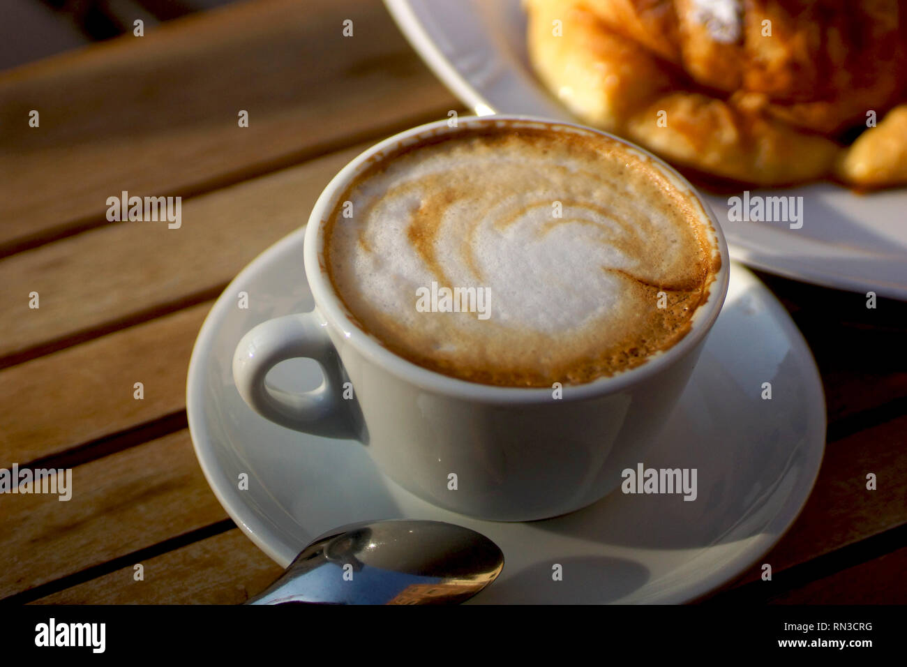 Köstlich heißen italienischen Cappuccino steht in eine Tasse mit Untertasse auf einem Holztisch. Im Hintergrund ein frisches Croissant auf einem Teller. Stockfoto
