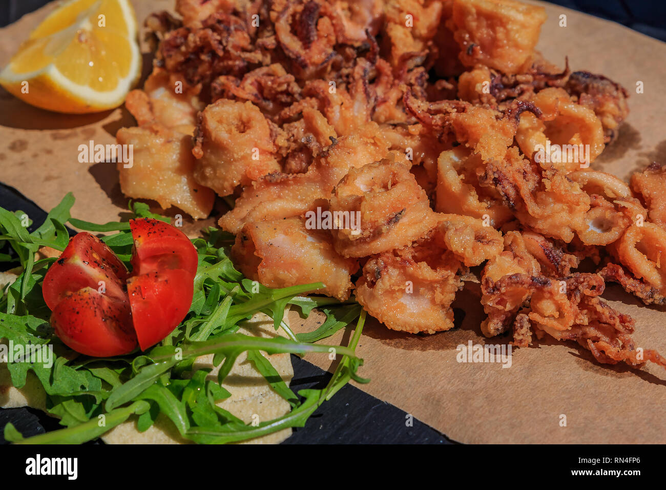 Knusprig frittierte Calamari Ringe und Tentakeln auf einem urigen Board auf ein Stück Papier mit Zitrone und Salat mit Rucola und Tomaten mit Olivenöl o serviert. Stockfoto