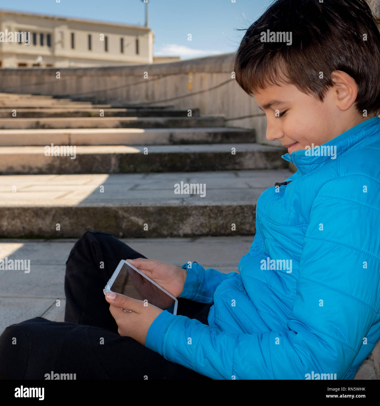 Ein Kind mit einer blau Daunenjacke, auf dem Steinboden sitzen und an die Wand gelehnt, spielt mit einem digitalen Tablet Stockfoto