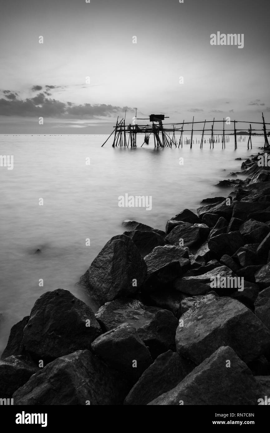 Holzsteg mit der felsigen Küste bei Sonnenuntergang. Lange Belichtung Vertikal Schwarz und Weiß Stockfoto