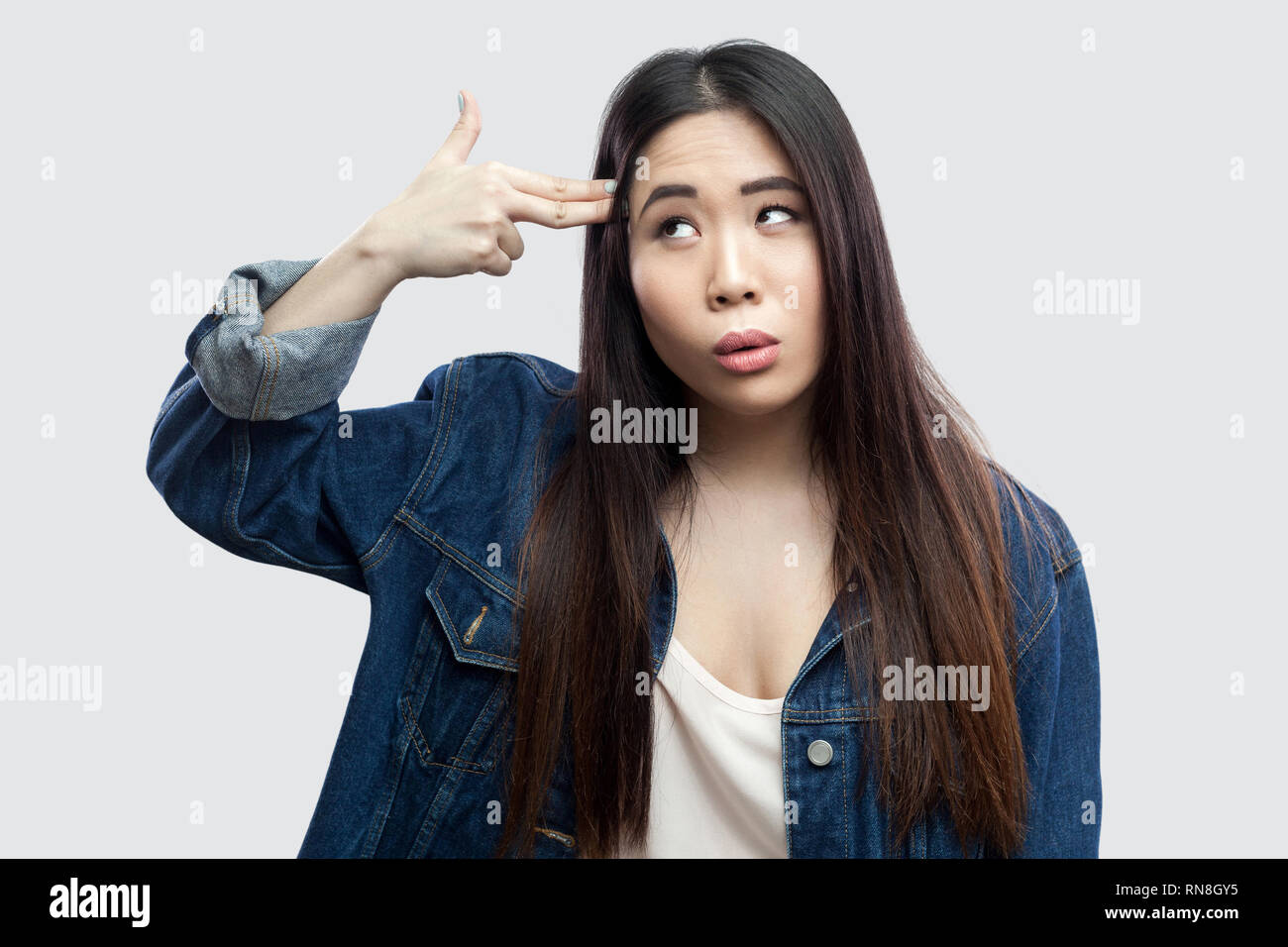 Portrait von einsamen gedrückt schöne Brünette asiatische junge Frau in lässigem Blue Denim Jacke mit Make-up stehend mit Gewehr Pistole am Kopf. indoor Stu Stockfoto