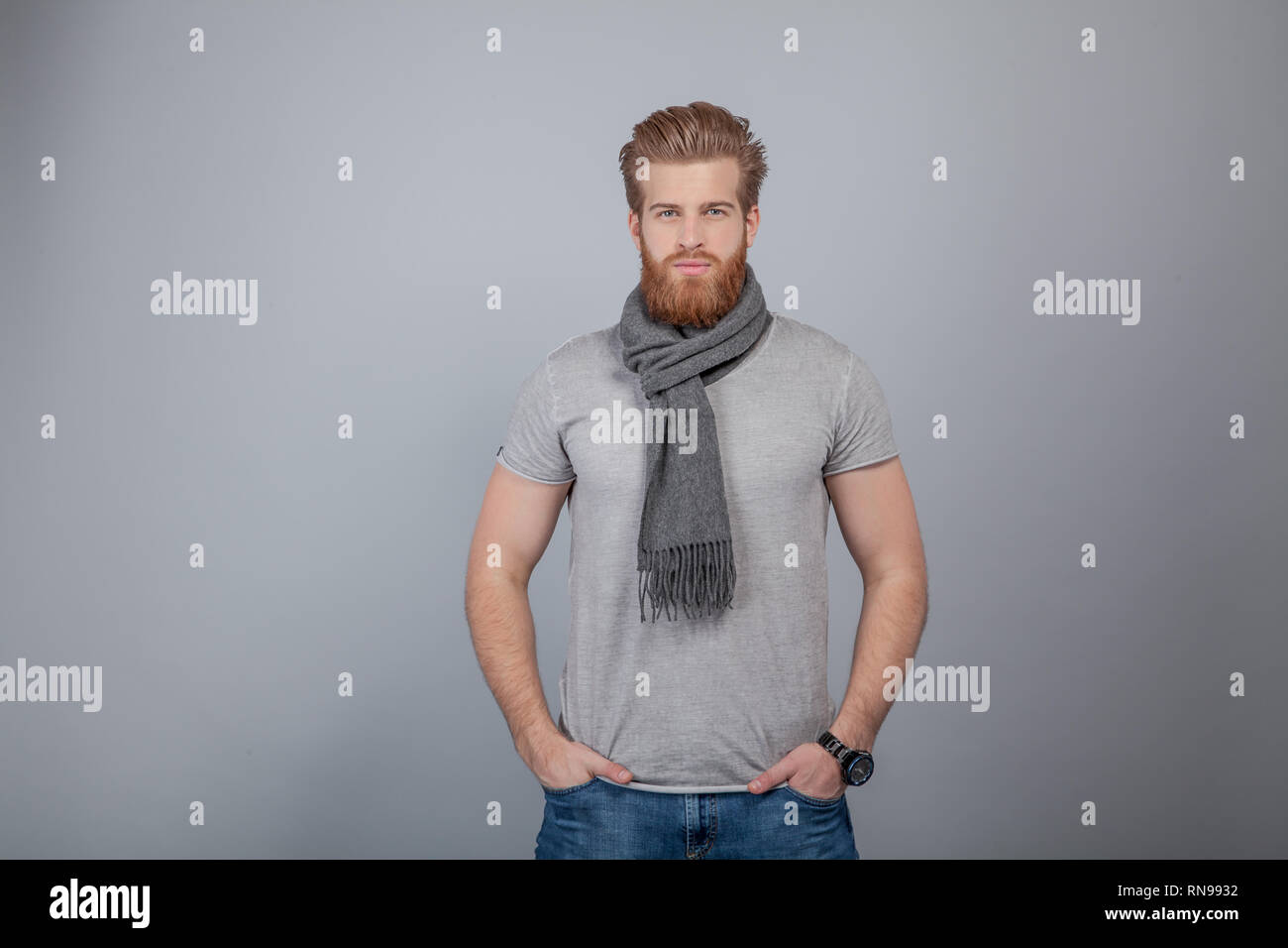 Die Jungen gut aussehenden bärtigen Mann, der durch die graue Wand Schal tragen, grau t-shirt und schaut direkt in die Kamera schaut. Stockfoto