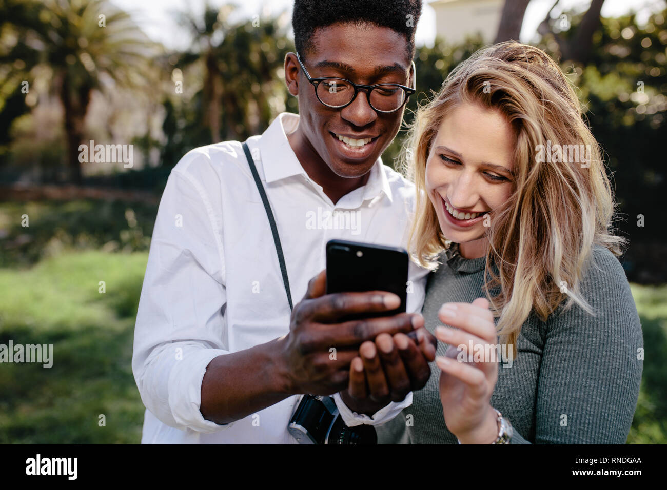 Lächelnd gemischtrassiges Paar stehen zusammen und Betrachten der Bilder auf dem Smartphone. Junger Mann und Frau an die Fotos auf Handy suchen Stockfoto