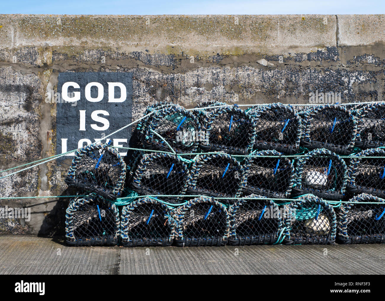 Creel Netze stapelten sich an einem sonnigen Tag mit einer "Gott ist die Liebe'-Schild am Hafen Wand hinter Ihnen, Gardenstown, Aberdeenshire lackiert Stockfoto