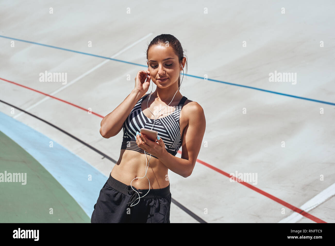 Junge Frau mit schwarzen Sportswear stehend auf Rennstrecke und Musik zu hören. Läuferin üben auf Athletik Rennstrecke. Sie ist glücklich und mit sich selbst zufrieden Stockfoto