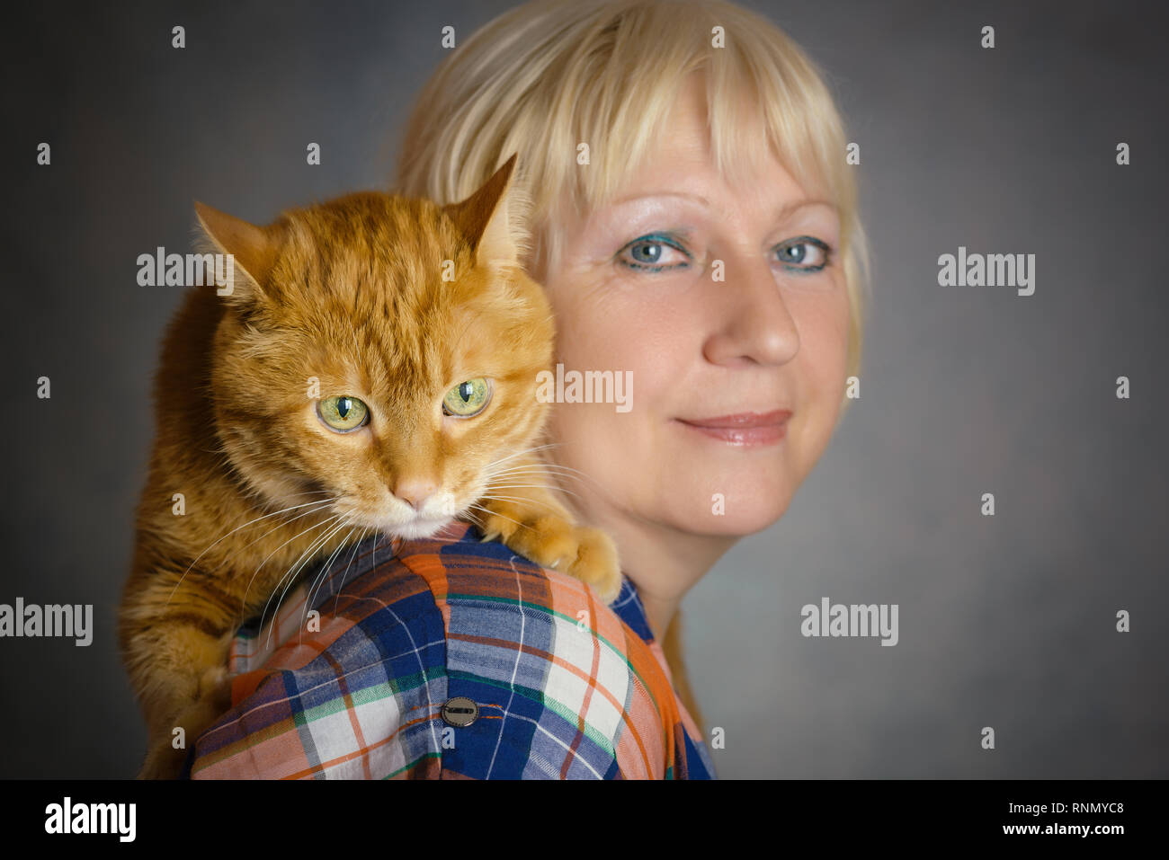Schön gealterte Frau umarmte ihre rote Katze auf grauem Hintergrund. Fokus auf eine Katze. Stockfoto