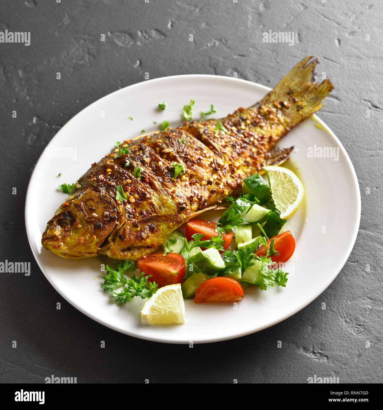 In der Nähe von Ofen gebackenen Fisch auf dem Teller über schwarzen Stein. Gesunde Meeresfrüchte. Lecker gekochten Fisch Salat von frischem Gemüse (Gurke, Tomate, Gr Stockfoto