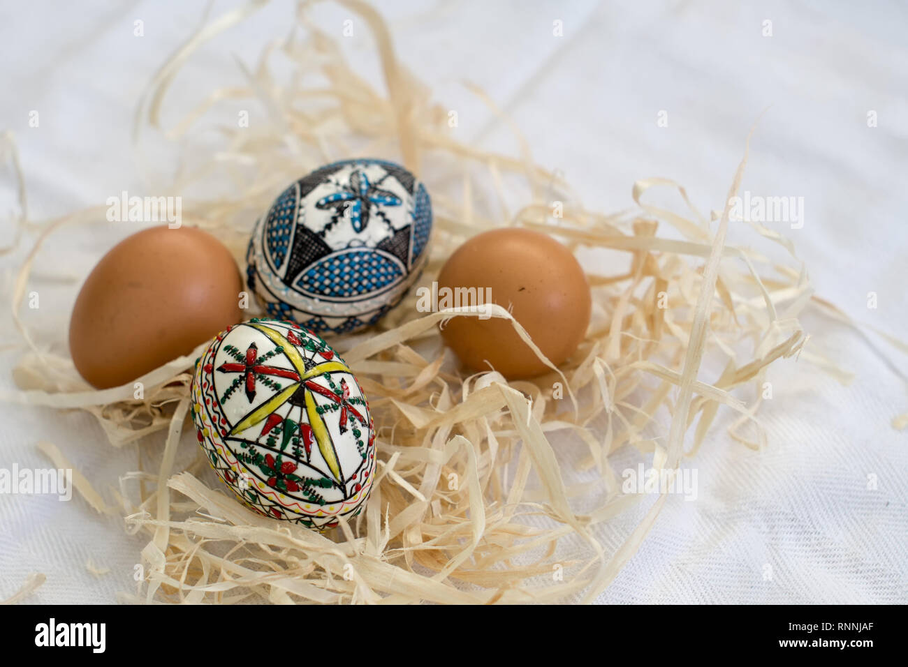 Traditionelle handgefertigte Ostereier im Stroh auf einem weißen Tuch und wenige Organische frische Eier im Hintergrund. Traditionelle Muster von Bucovina Stockfoto