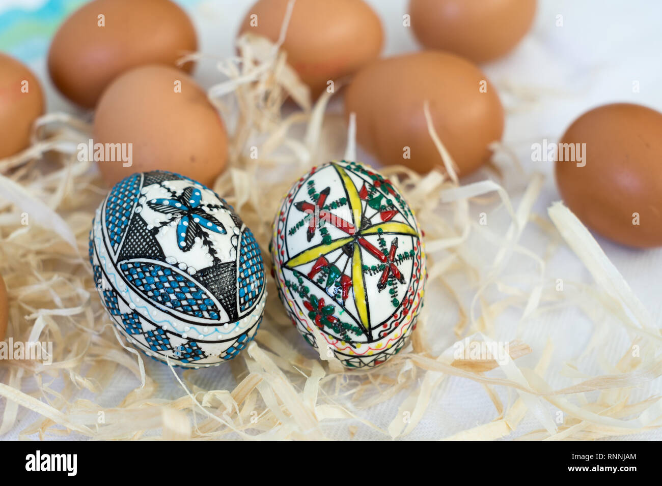 Traditionelle handgefertigte Ostereier im Stroh auf einem weißen Tuch und wenige Organische frische Eier im Hintergrund. Traditionelle Muster von Bucovina Stockfoto