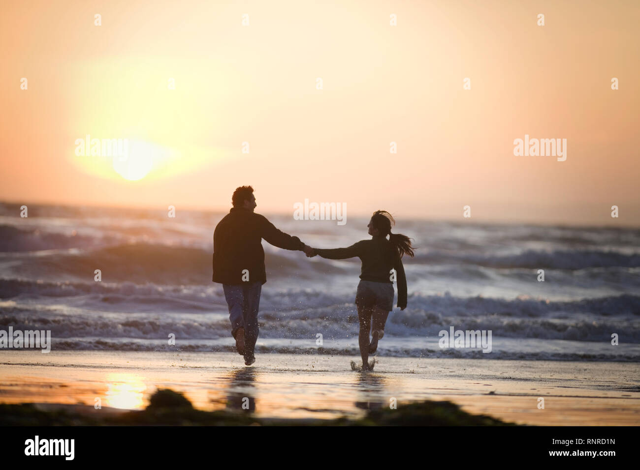 Junge erwachsene Paare Hand in Hand in Richtung der Brandung am Strand bei Sonnenuntergang. Stockfoto