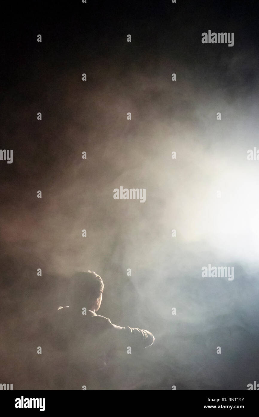 Ein junger Mensch, der ein geheimnisvolles helles Licht betrachtet, das Wolken aus Rauch oder Nebel im Dunkeln beleuchtet, geeignet für einen Bucheinband Stockfoto