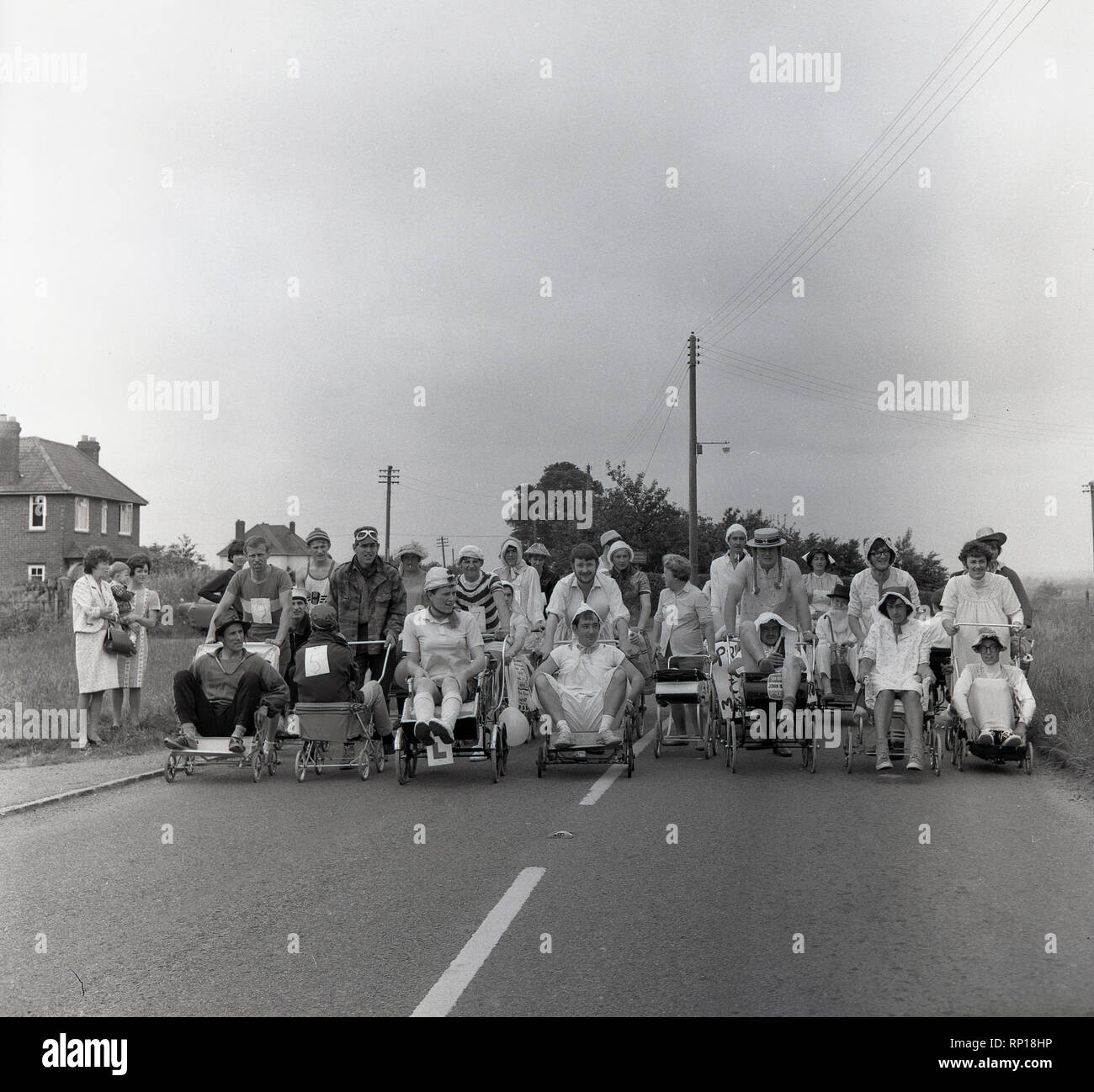 1965, historische, männlichen Konkurrenten in einem englischen Dorf pram Rennen, das Tragen von Fancy Dress oder Kostüme, Line-up mit ihren Fahrgästen und Kinderwagen auf der Landstraße für den Beginn der Veranstaltung, England, UK. Stockfoto