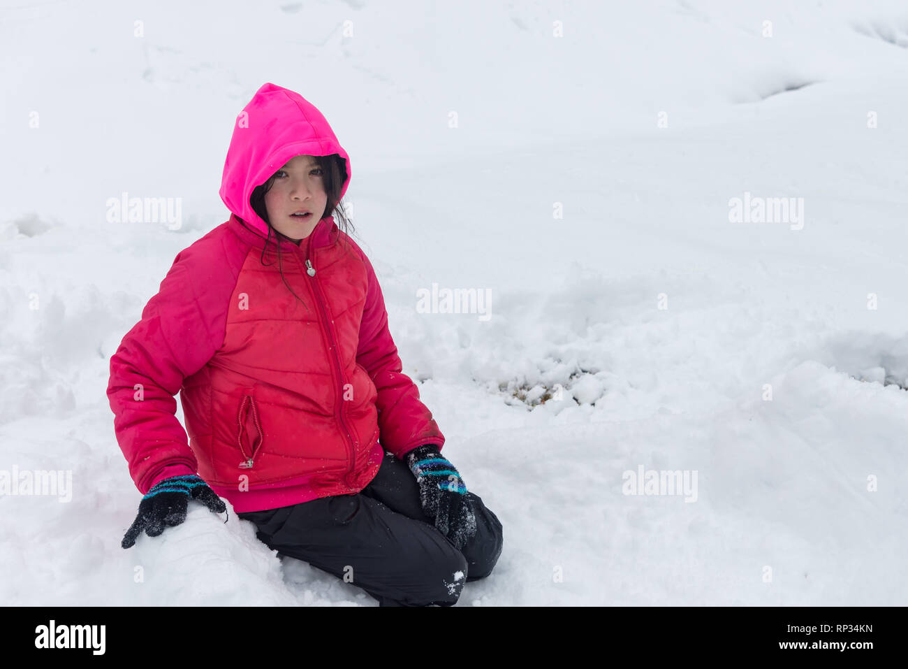 Junge Mädchen mit rosa Haube und Jacke und Handschuhe kniend im Schnee Stockfoto